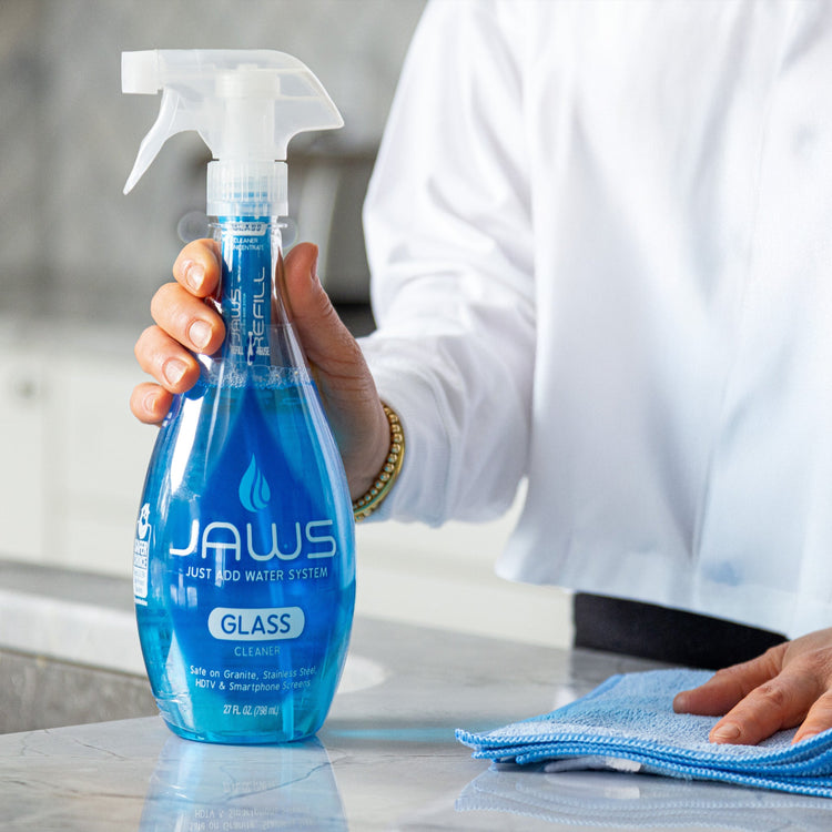 Person holding a blue glass cleaner bottle labeled 'JAWS' on a countertop.