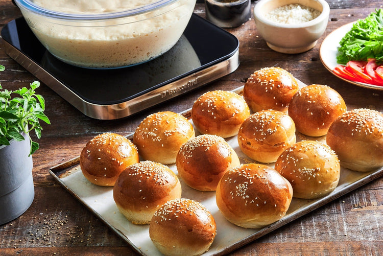 Baked buns on a baking tray with a bowl of dough and ingredients in the background.