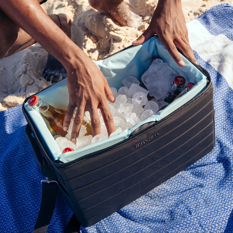 Person opening a cooler box filled with ice and drinks on a sandy beach.