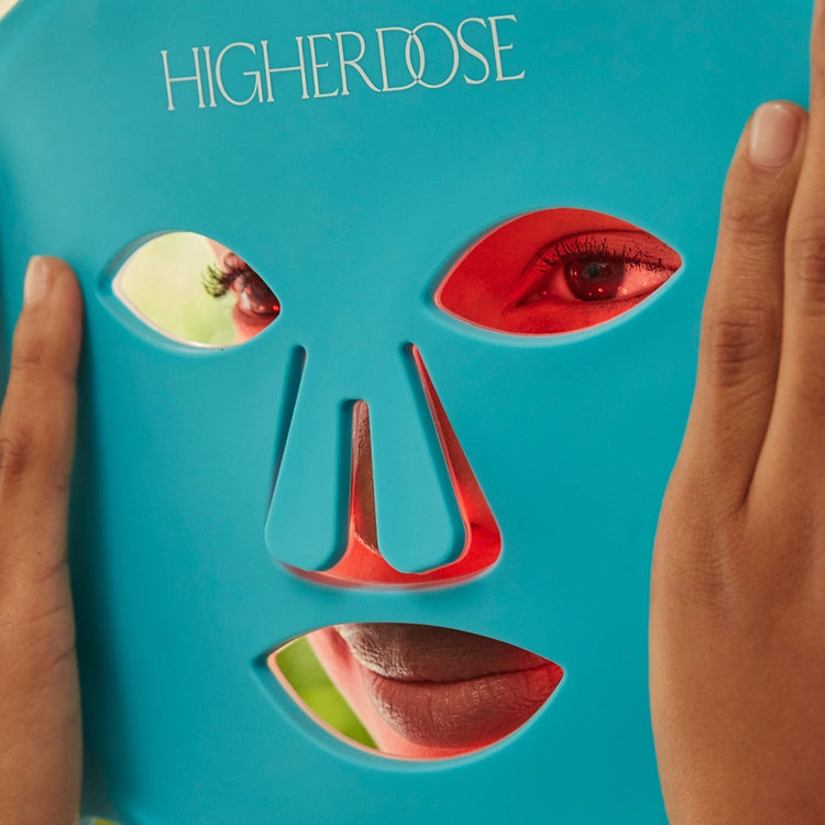 Blue sheet mask with face design held by hands against a blurred green background