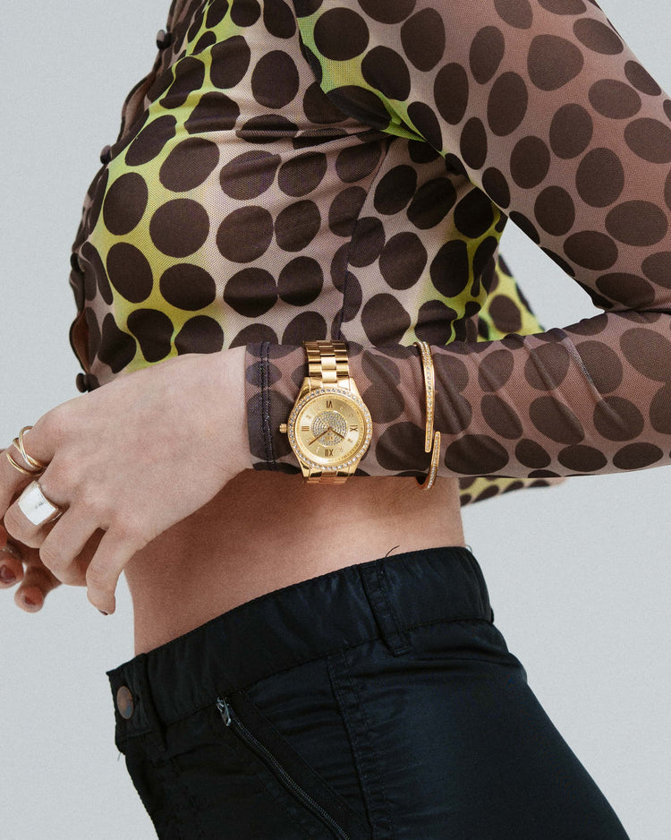 Gold watch on a wrist with a patterned sleeve and black pants against a neutral background