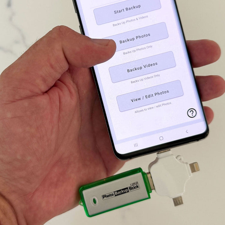 Hand holding a smartphone with photo backup app open, connected to a Photo Backup Stick.