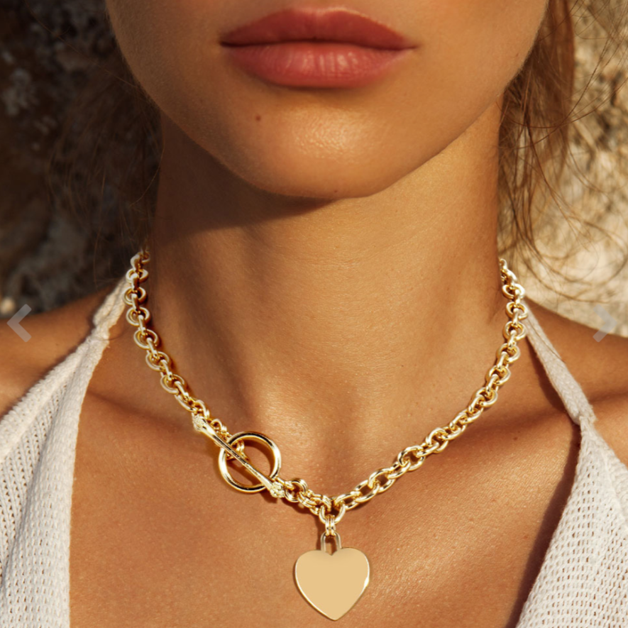 A close-up image of a woman wearing a gold toggle necklace with a heart pendant.