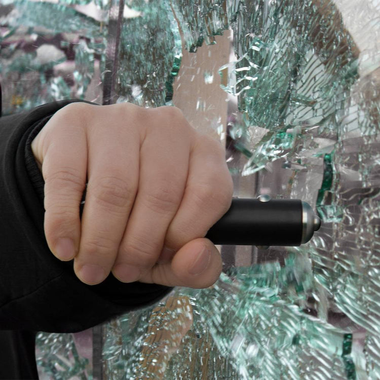 A hand holding a black React 7-in-1 vehicle emergency multi-tool with a seat belt cutter on the end, shown against a background of shattered glass.