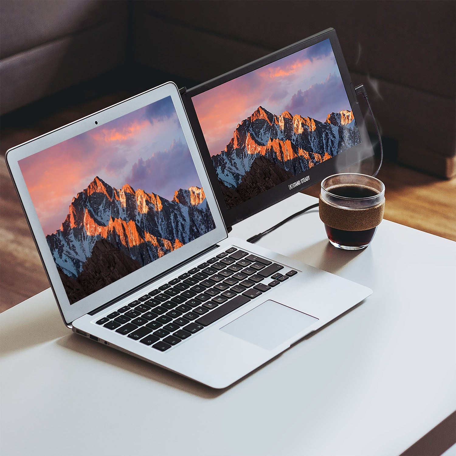 Two laptops displaying mountain scenery on a table with a steaming cup of coffee.