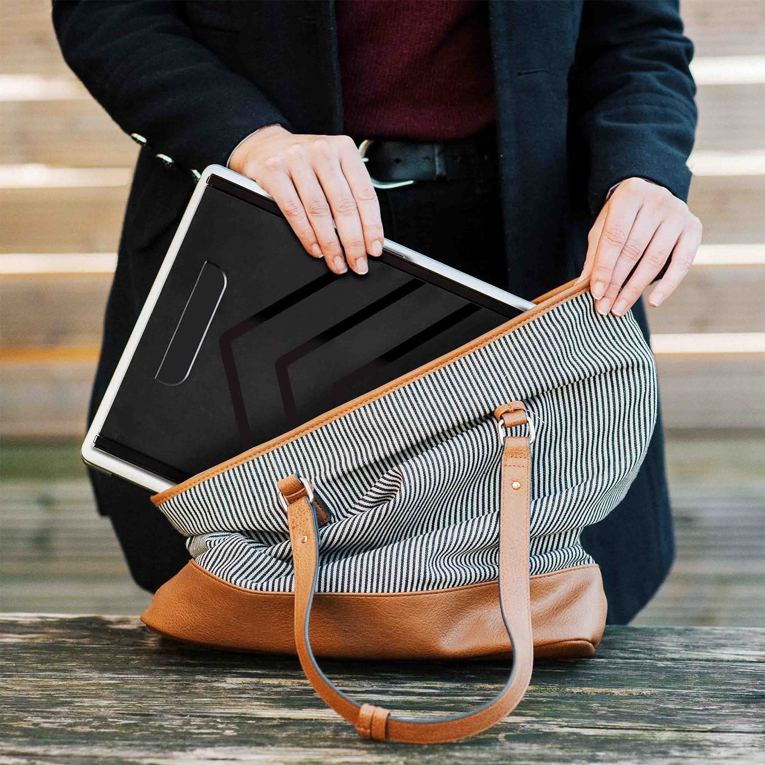 Person holding a black laptop in a striped bag with a brown strap.