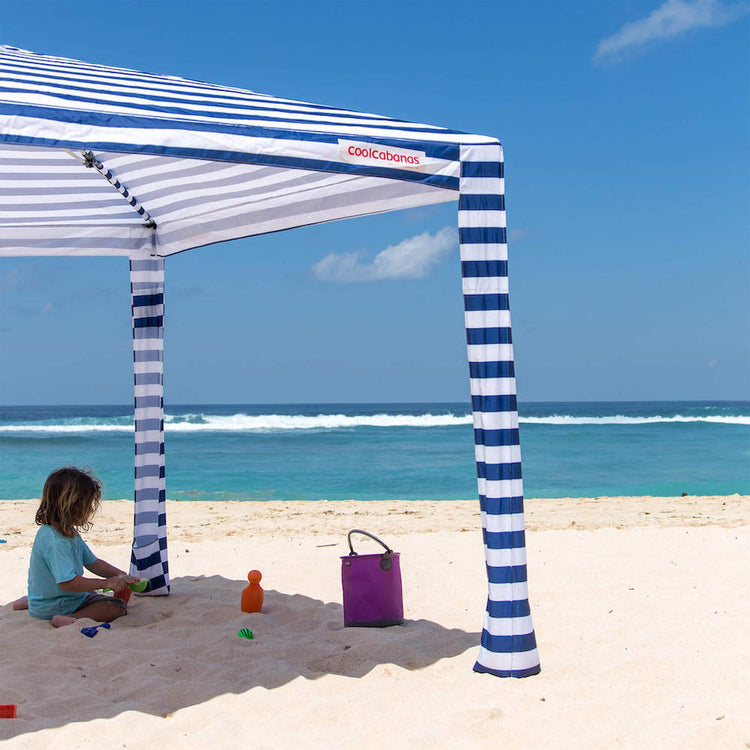Child playing under a blue and white striped canopy on a sandy beach with ocean view.