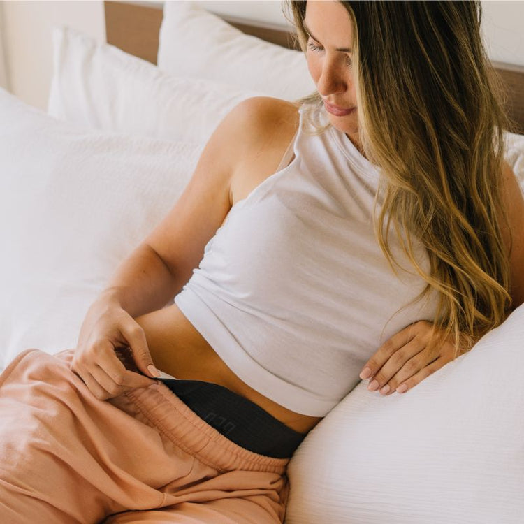 Woman sitting on a bed wearing a white tank top and brown pants Using GEO device