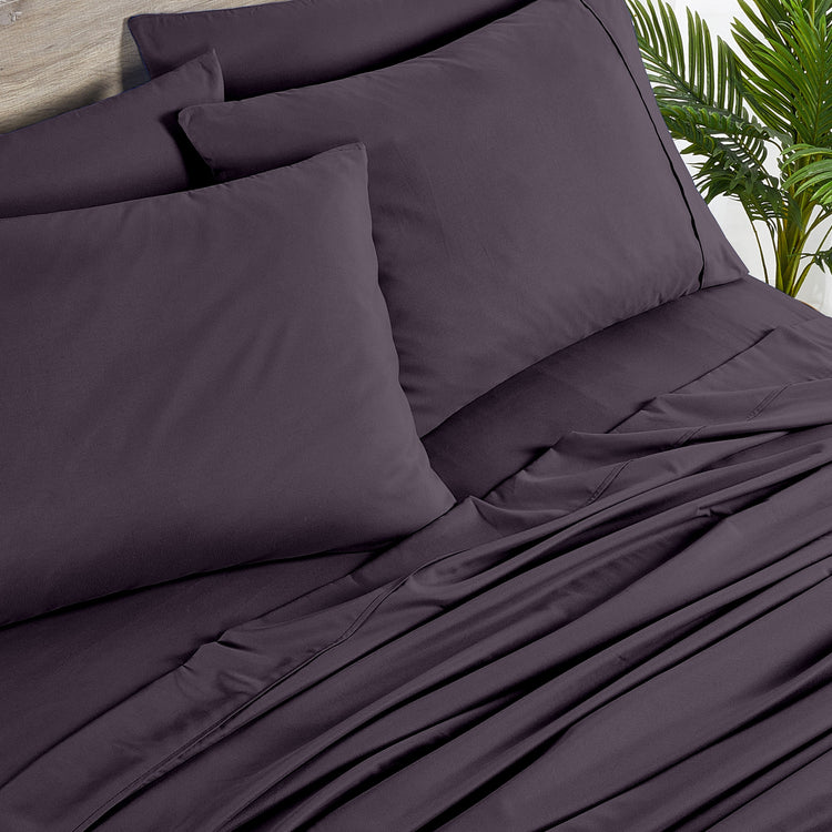 Purple bedding set with pillows on a bed, with a plant in the background.