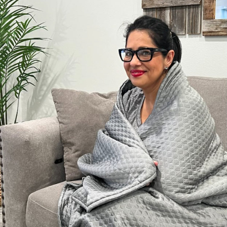 Woman sitting on a couch wrapped in a gray weighted blanket, with a plant and wooden wall in the background.