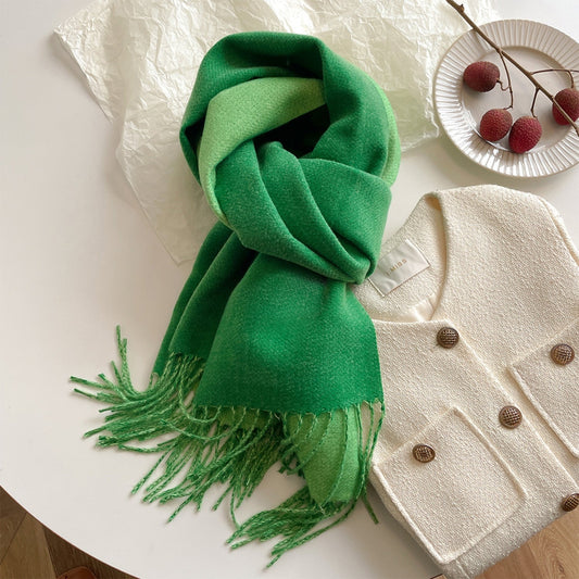 A green reversible pashmina scarf with fringe detailing, displayed on a white surface next to a beige coat and a pretzel.
