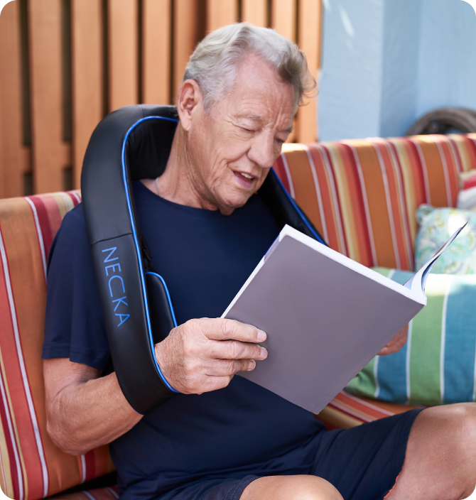 Man sitting on a striped couch wearing a neck massager and reading a book.