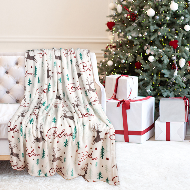 Christmas-themed blanket with reindeer and 'Believe' text draped over a chair, with a decorated Christmas tree and presents in the background.