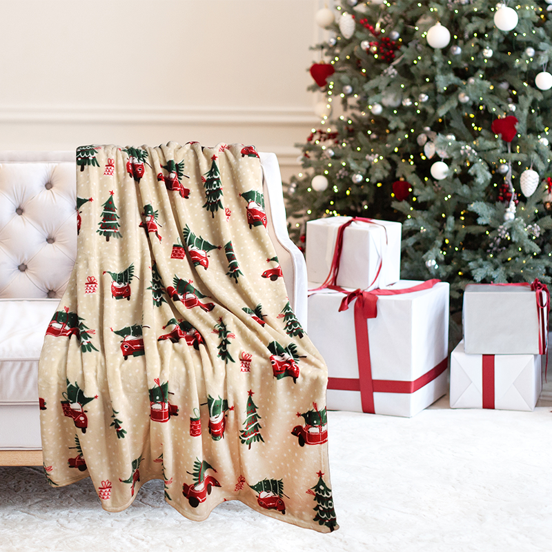 Christmas-themed blanket with festive design draped over a chair next to wrapped gifts and a decorated tree.