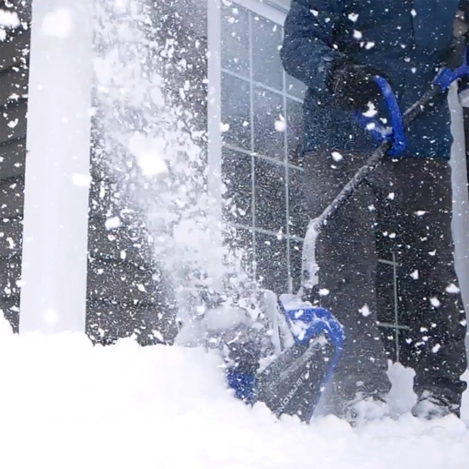 Person using a snow blower to clear snow outside a building during a snowstorm.