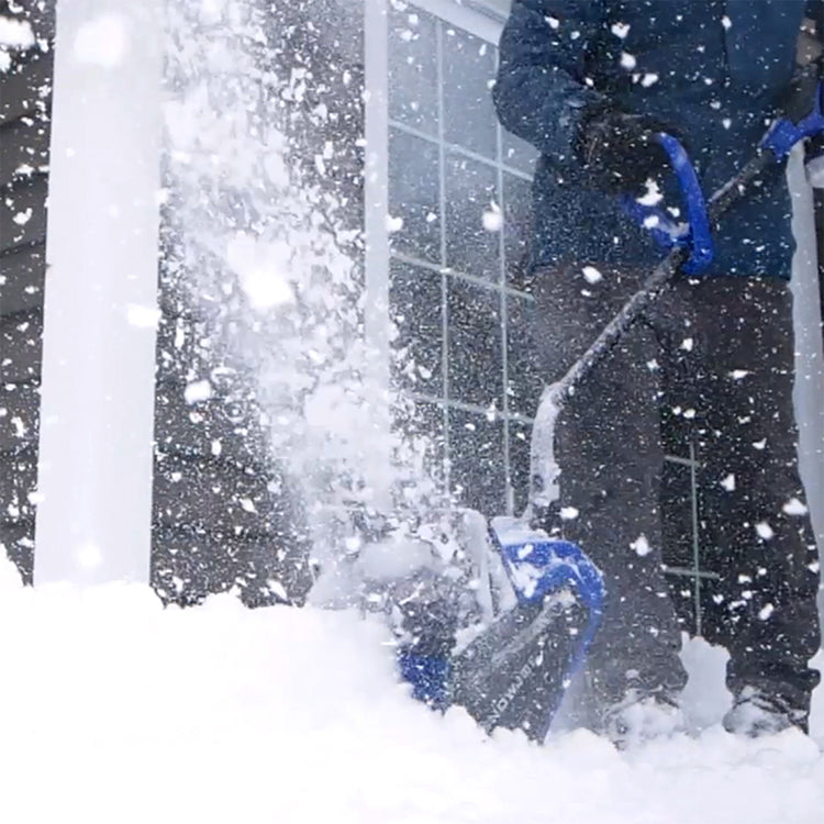 Person using a snow blower to clear snow outside a building during a snowstorm.