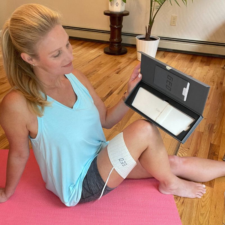 Woman sitting on a pink mat with a device labeled 'GEO' on her leg, in a home setting.