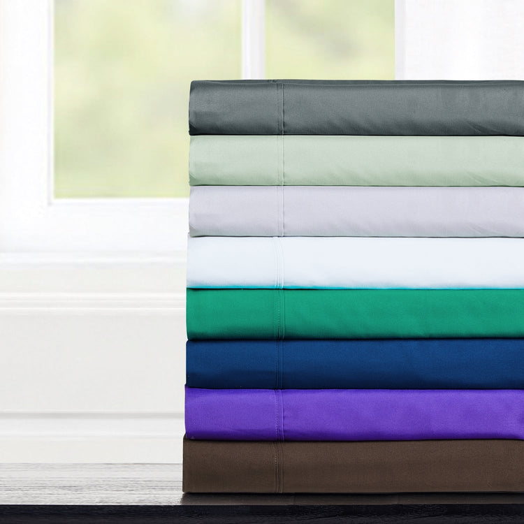 A stack of folded bamboo bed sheets in various colors including white, green, blue, purple, and brown, displayed in front of a window.
