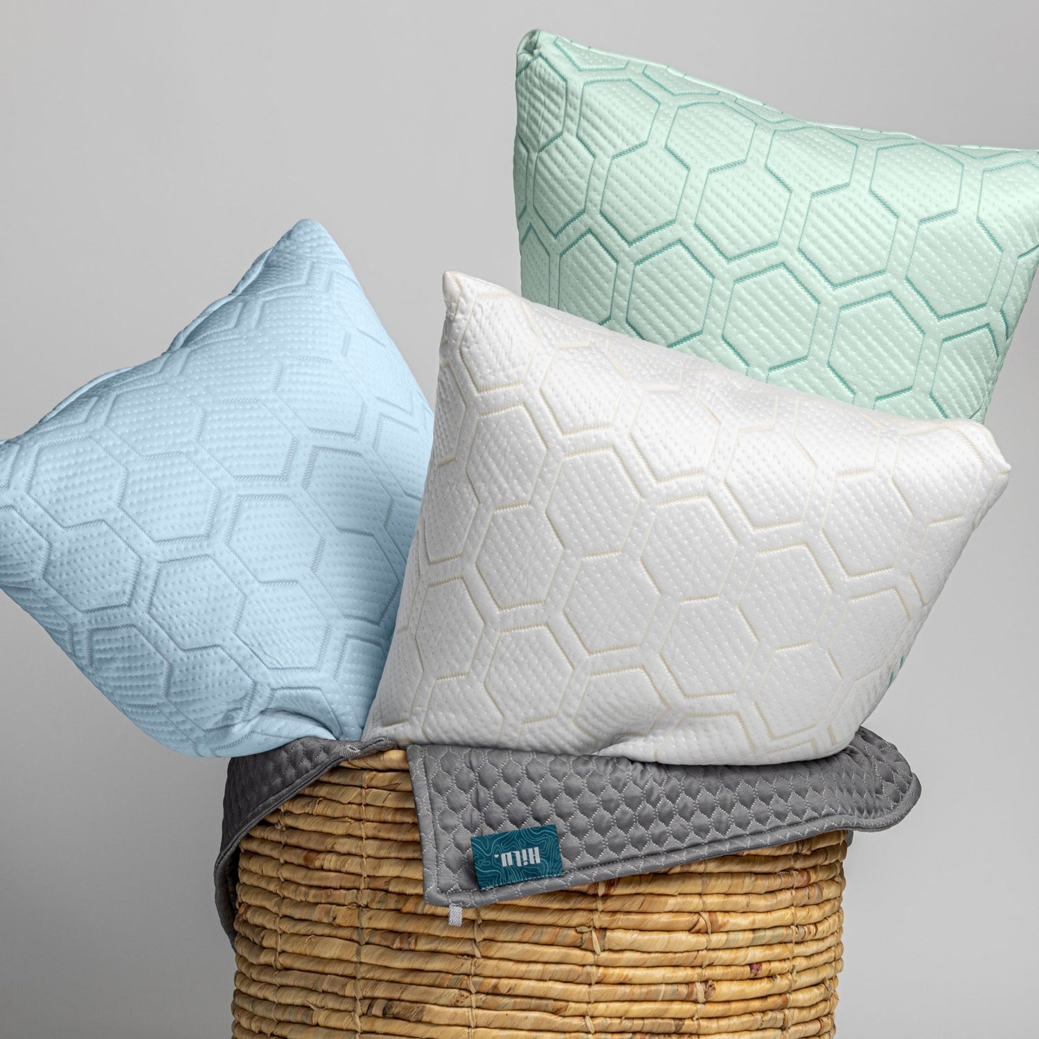 Three textured pillows in blue, white, and green on a wicker basket with a gray background.
