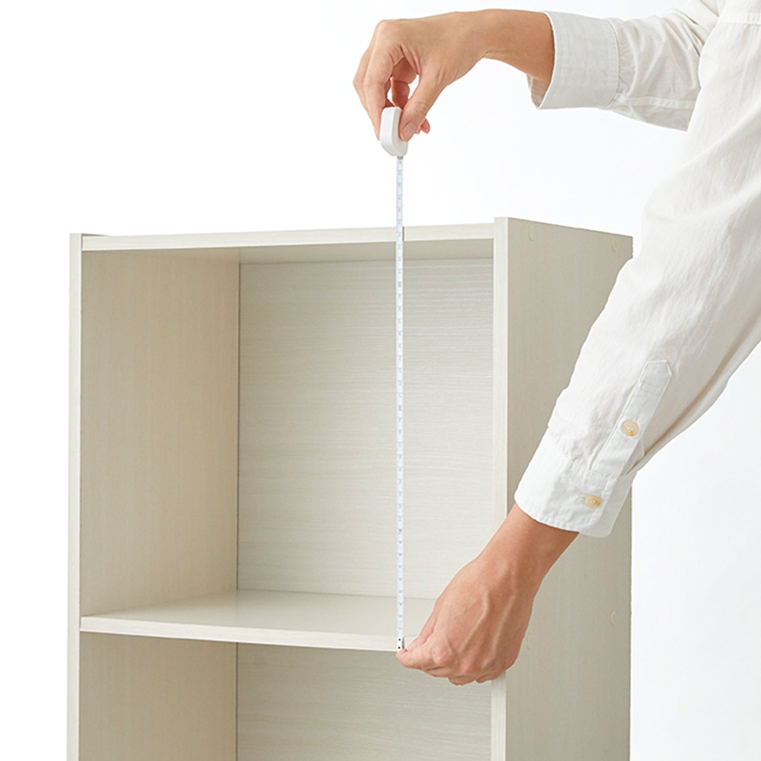 A person in a white shirt uses a white tape measure to determine the height of a light wood two-shelf bookcase against a white background.