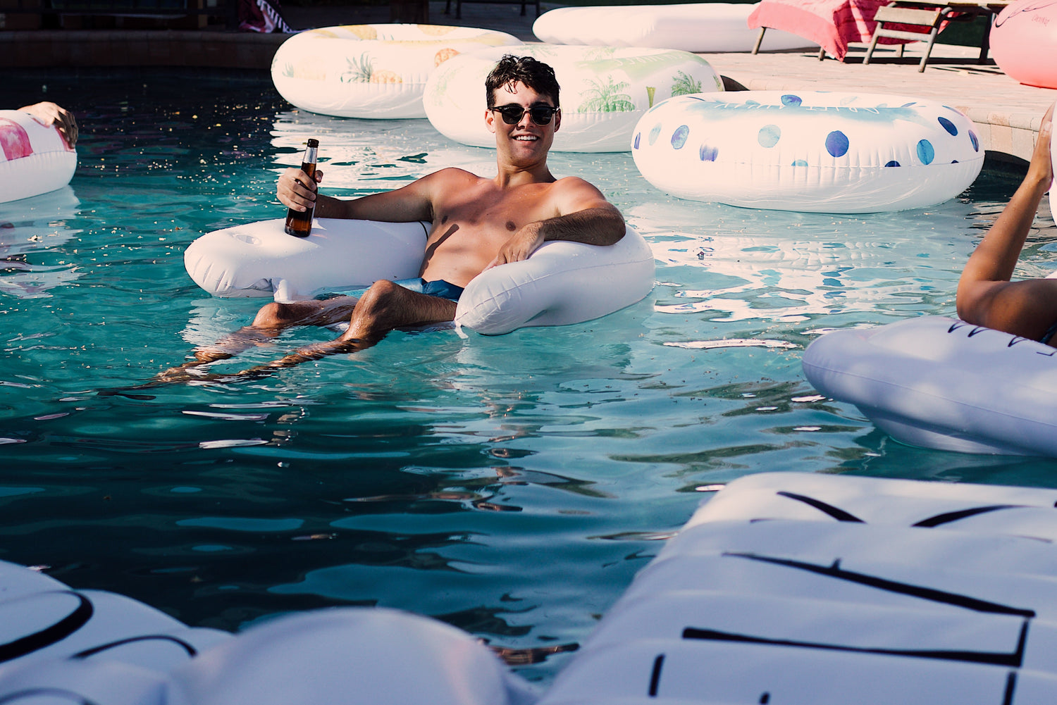Man relaxing on a inflatable pool float with drinks, surrounded by other pool-goers and inflatables.