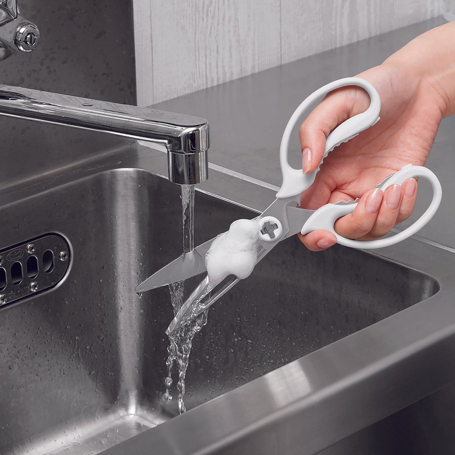 A hand holds a pair of kitchen shears with white handles and a white rubber blade protector over running water in a stainless steel sink.
