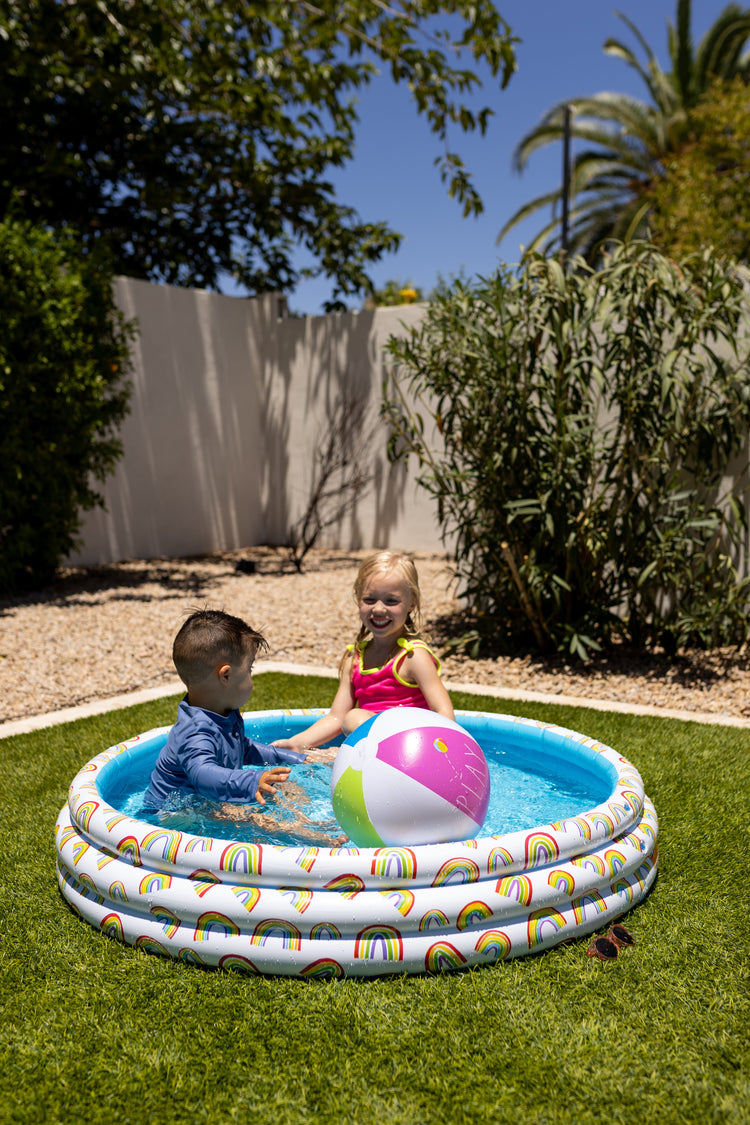 Two children playing in a small inflatable pool with a colorful ball in a backyard.