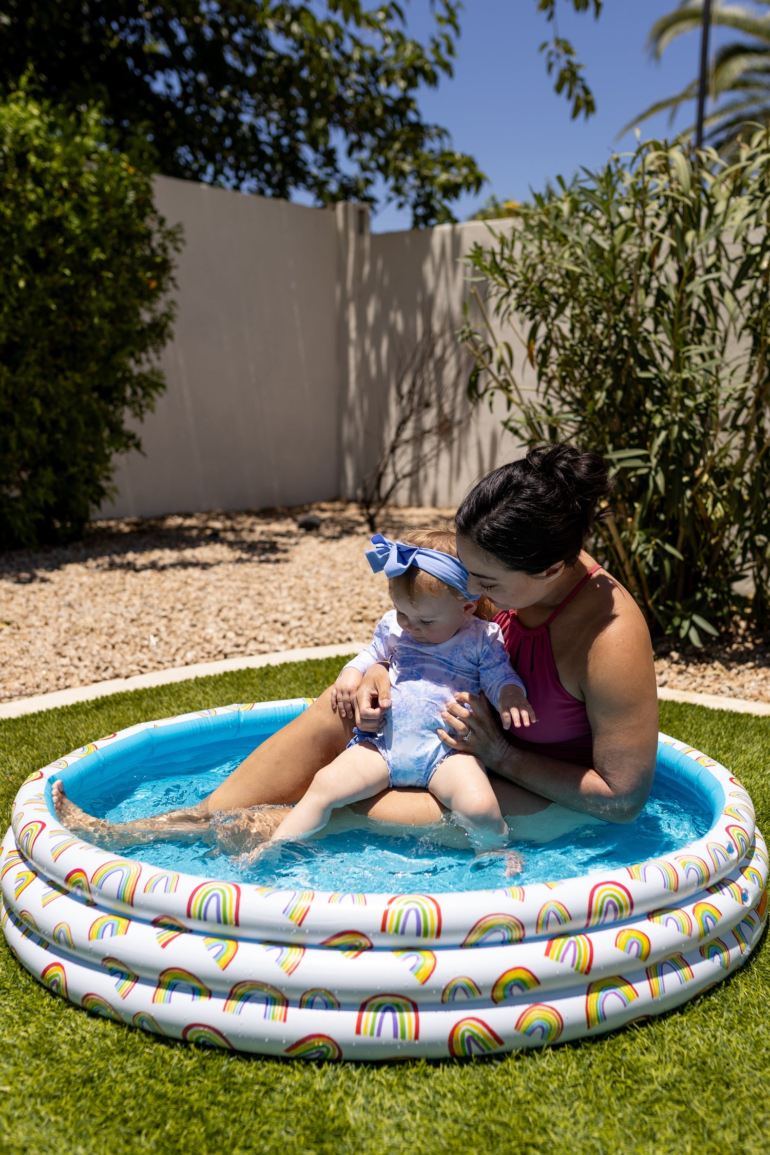 Woman and child in a small inflatable pool with rainbow design on grass.