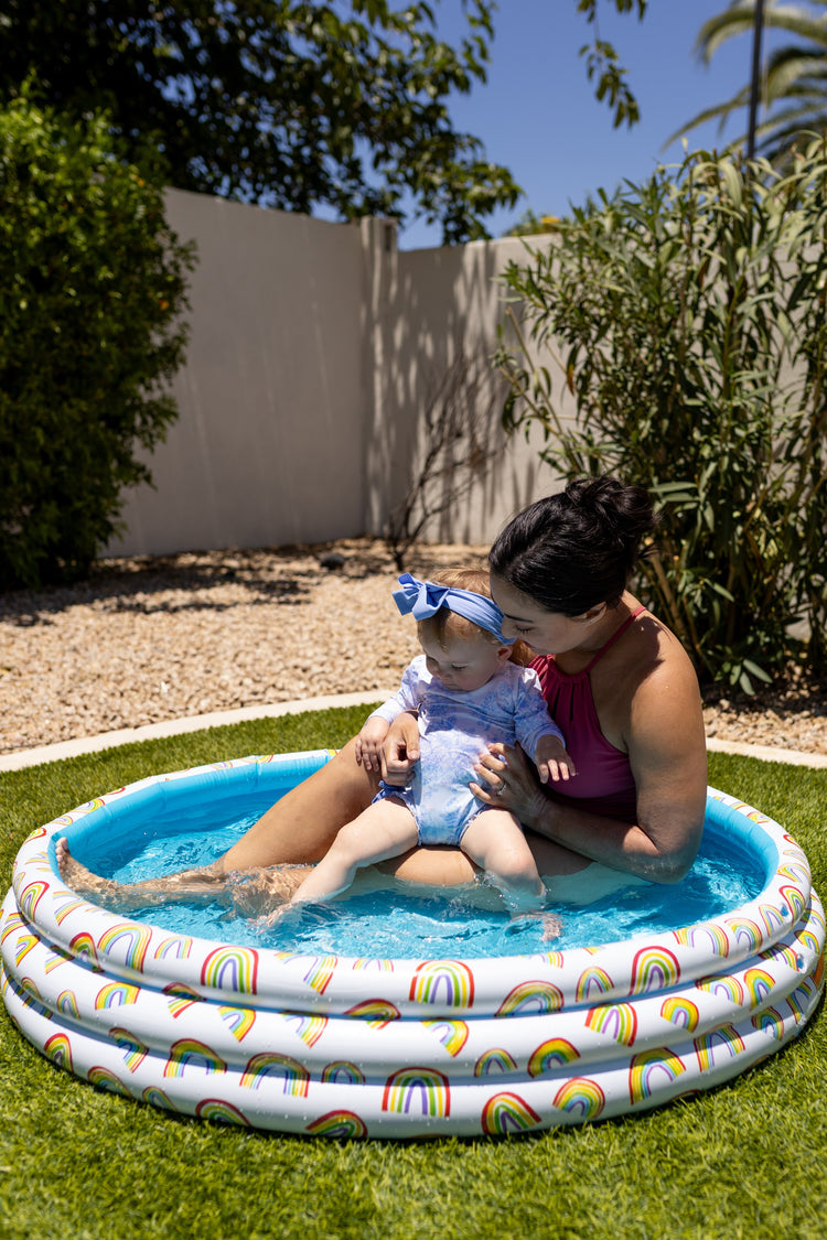 Woman and child in a small inflatable pool with rainbow design on grass.