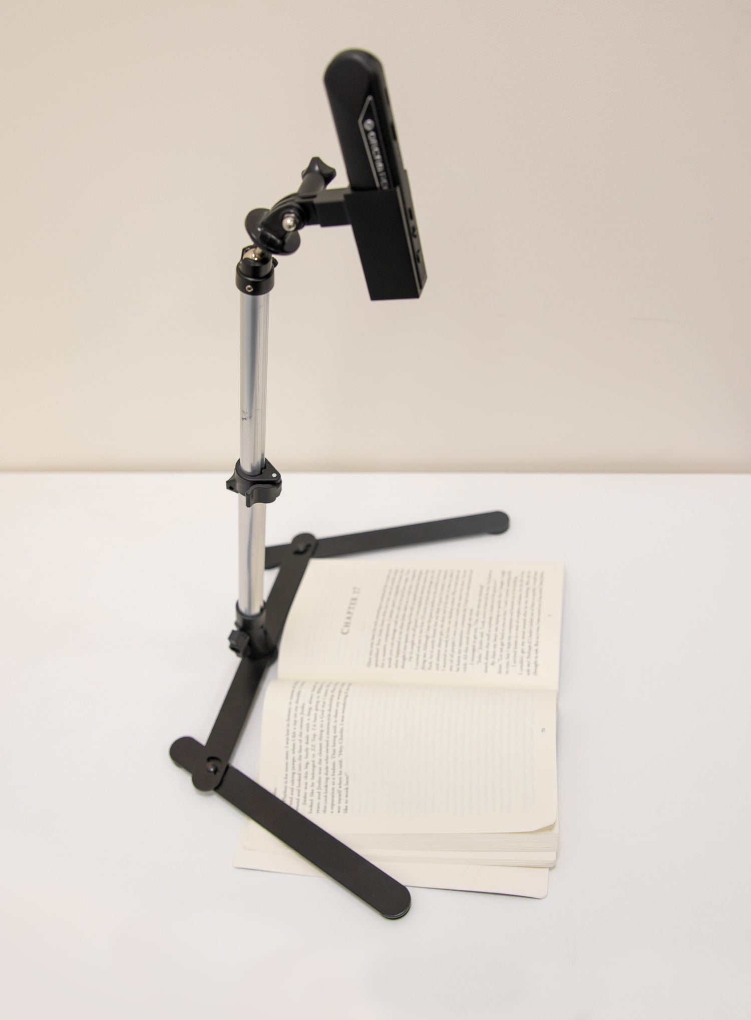 Black adjustable stand holding a tablet above an open book on a white surface.