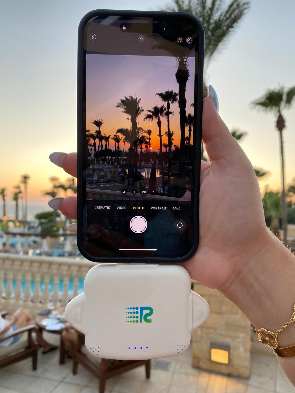 A hand holds a smartphone in front of a white portable projector dock, with a resort poolside patio and palm trees visible in the background at sunset.