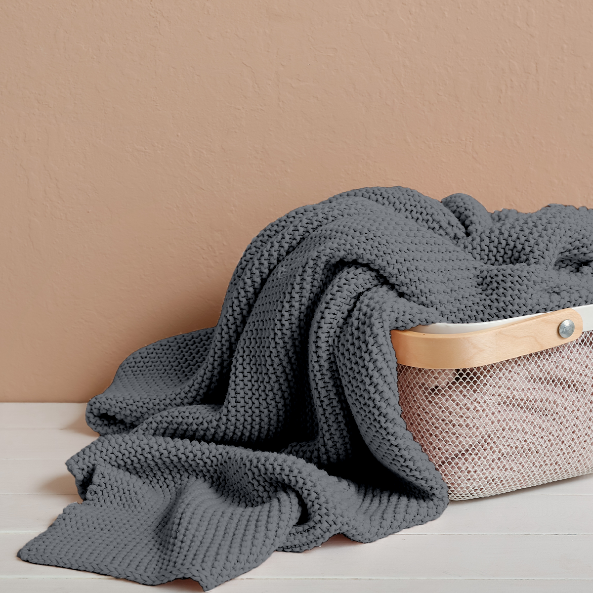 Gray knitted blanket draped over a beige woven basket against a beige wall.