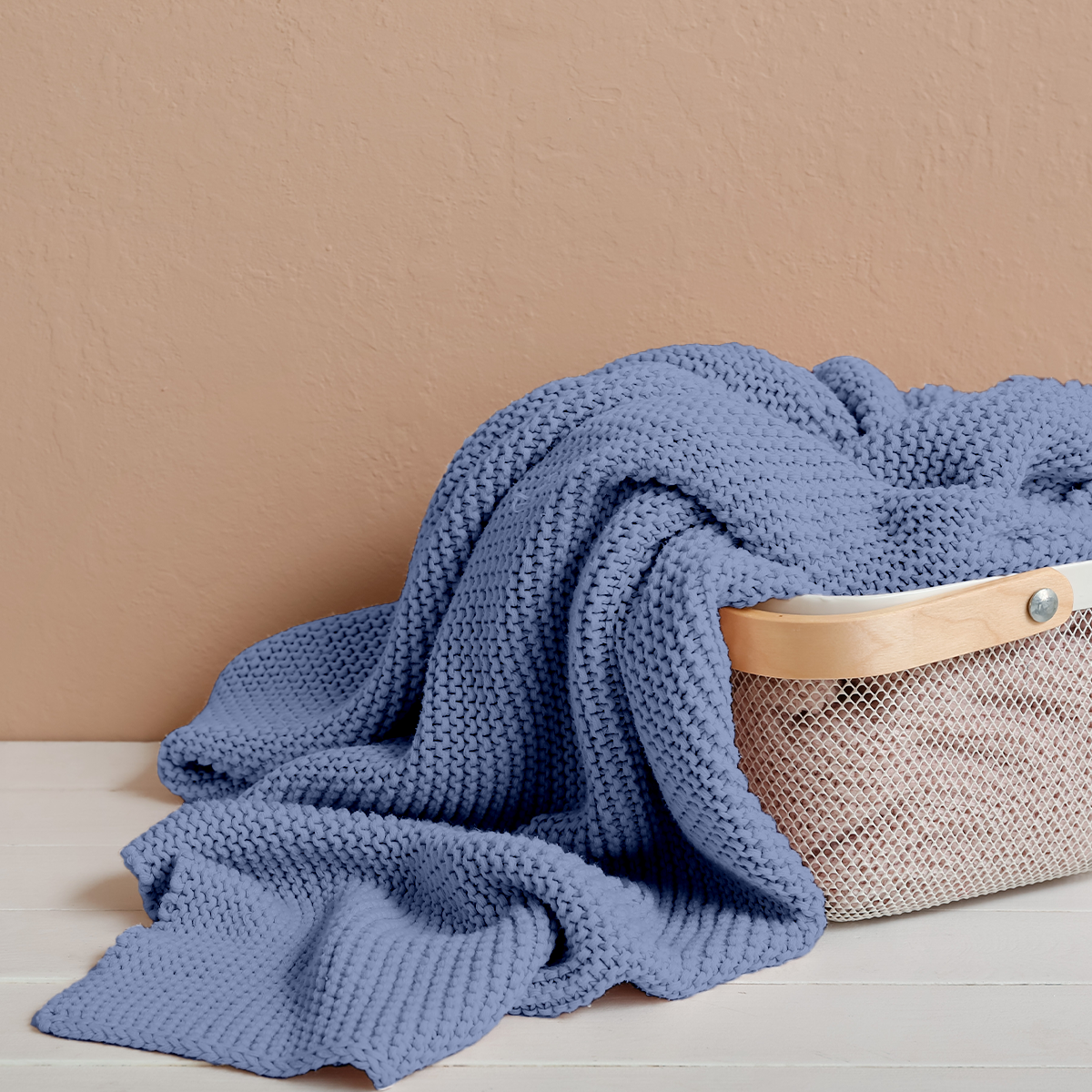 Blue knitted blanket draped over a woven basket on a beige surface with a beige wall background