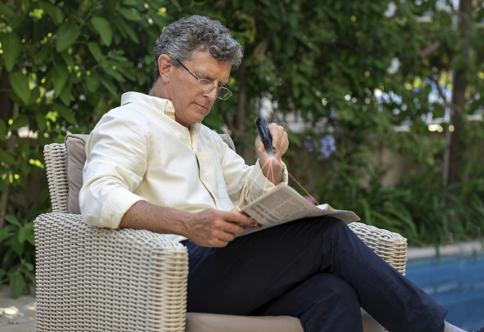 Man reading a newspaper outdoors in a wicker chair with greenery in the background