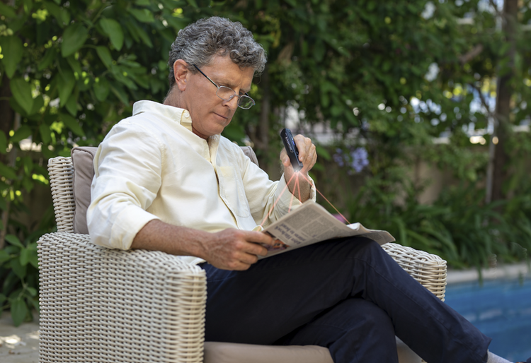 Man reading a newspaper outdoors in a wicker chair with greenery in the background