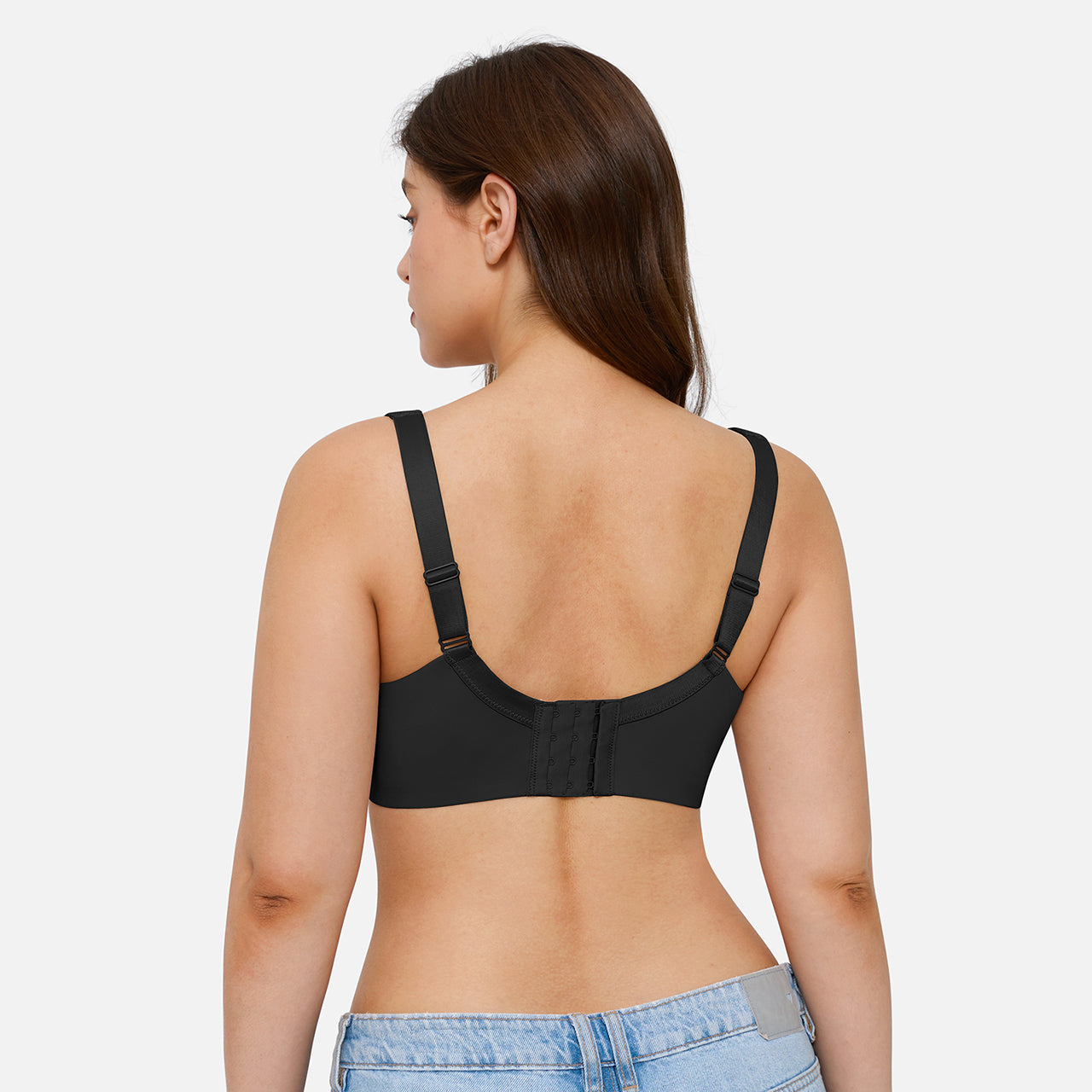 A woman shown from the back wearing a black bra with adjustable shoulder straps and a center back closure, paired with light blue jeans against a light gray studio background.