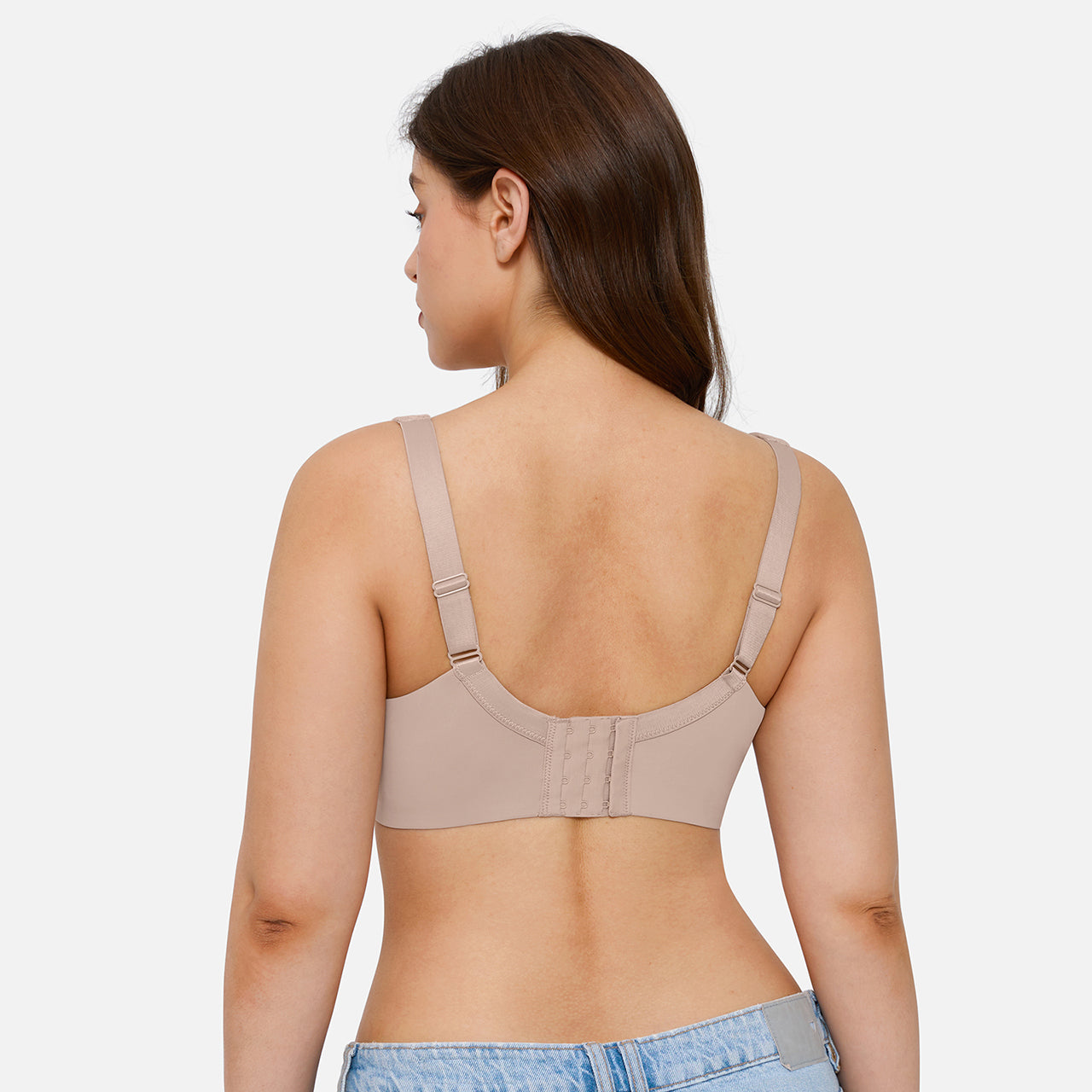 A woman shown from the back wearing a beige wireless bra with adjustable shoulder straps and a center gore closure, paired with light blue denim jeans against a light gray studio background.