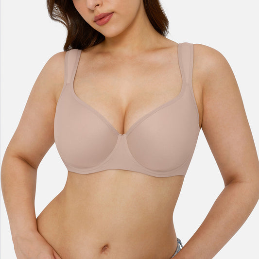 A woman modeling a seamless, wireless, scoop-neck bra in a light taupe-beige color, paired with light-wash blue jeans against a solid light gray background.