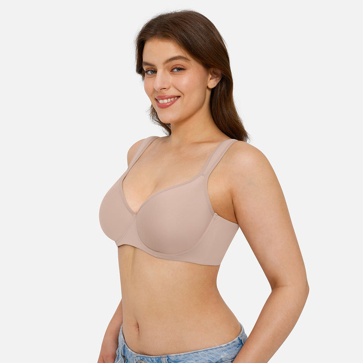 A woman modeling a seamless, underwire bra in a light taupe-beige color, paired with light-wash denim jeans against a solid light gray background.