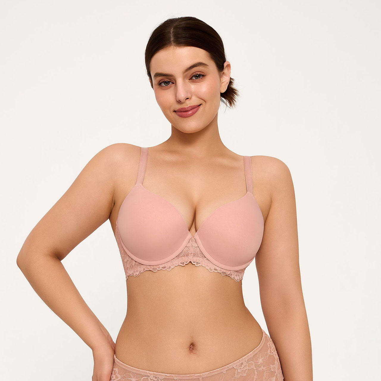 A woman modeling a light pink underwire bra with lace trim, paired with matching lace panties, against a plain light background.