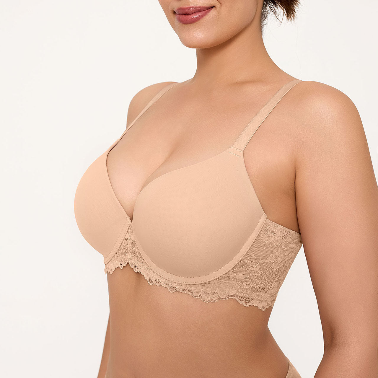 A close-up of a woman wearing a nude-beige wireless bra with delicate floral lace trim along the underband, photographed against a clean off-white studio background.