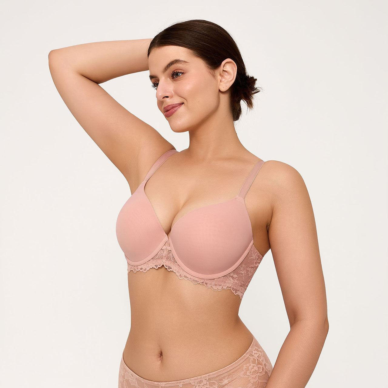 A woman modeling a light pink seamless bra with delicate lace trim and matching lace panties against a plain light gray background.