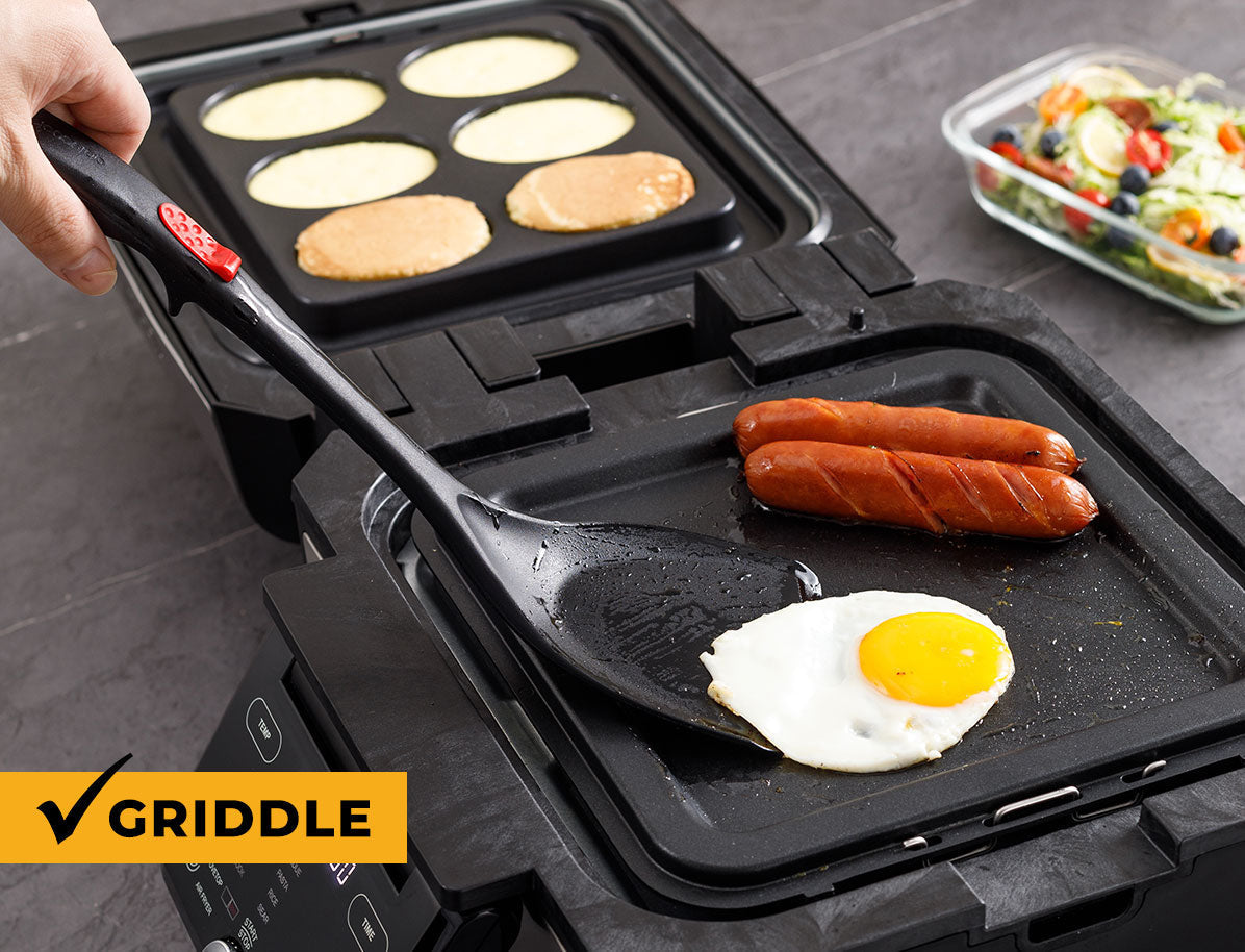 Multifunctional griddle with cooking surfaces, including pancakes, sausages, and an egg, on a gray surface.
