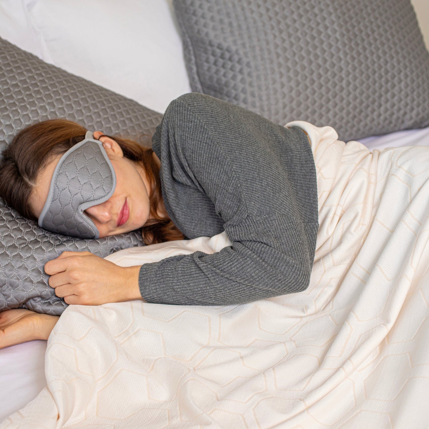 Person wearing a gray sleep mask lying in bed with white bedding.
