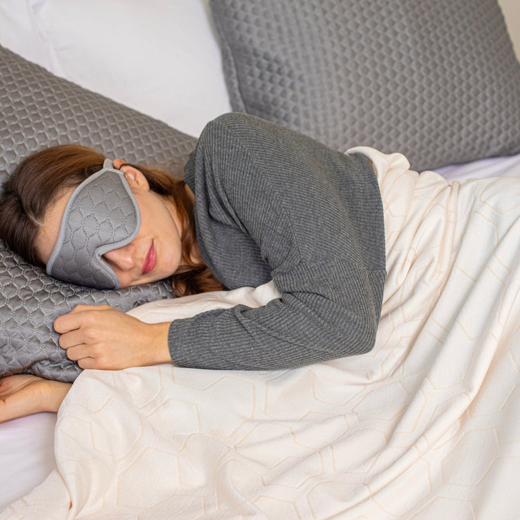 Person wearing a gray sleep mask lying in bed with white bedding.