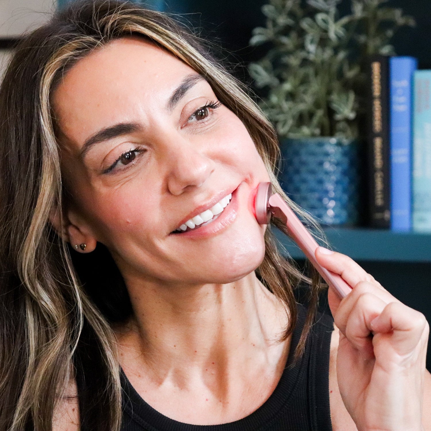 Woman applying lipstick in front of a bookshelf with books and a plant.