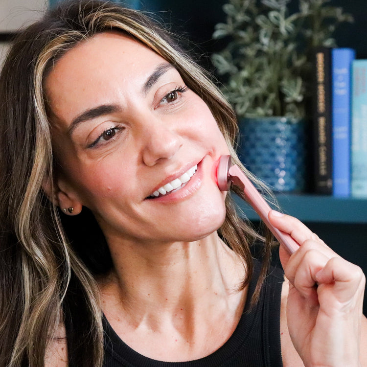 Woman applying lipstick in front of a bookshelf with books and a plant.