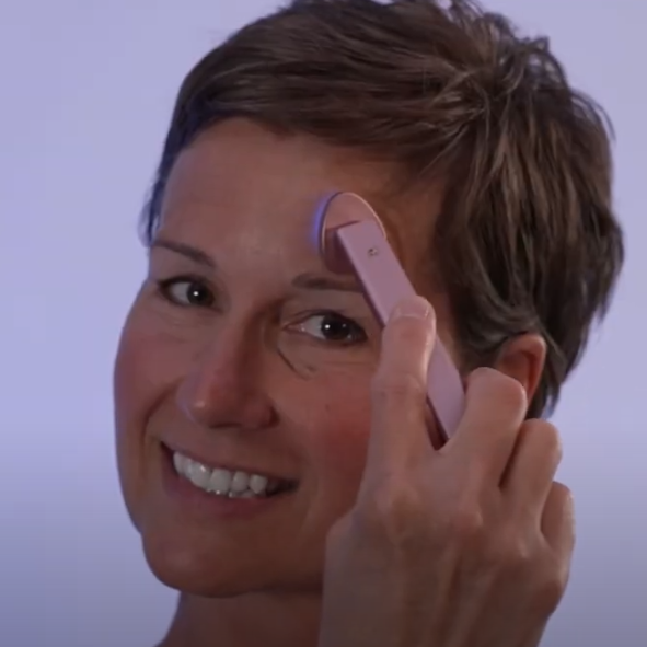 Woman using a pink skincare device on her face against a light purple background
