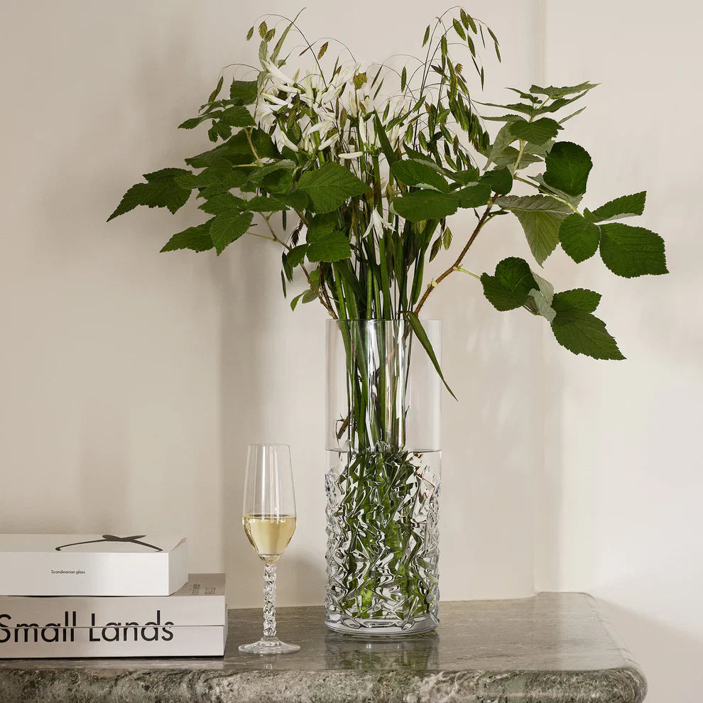 Clear glass vase with green leaves on a table next to a wine glass and books.