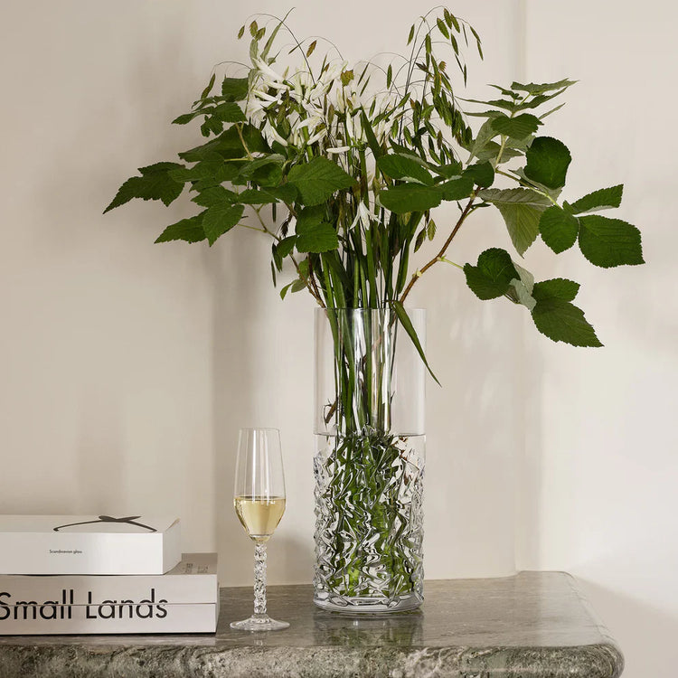 Clear glass vase with green leaves on a table next to a wine glass and books.